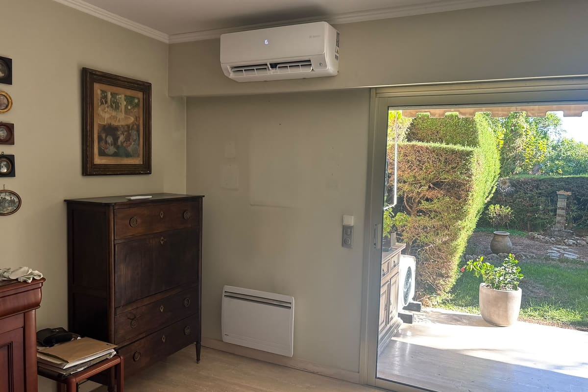 Climatiseur mural et radiateur électrique installés dans une pièce, avec vue sur le jardin.